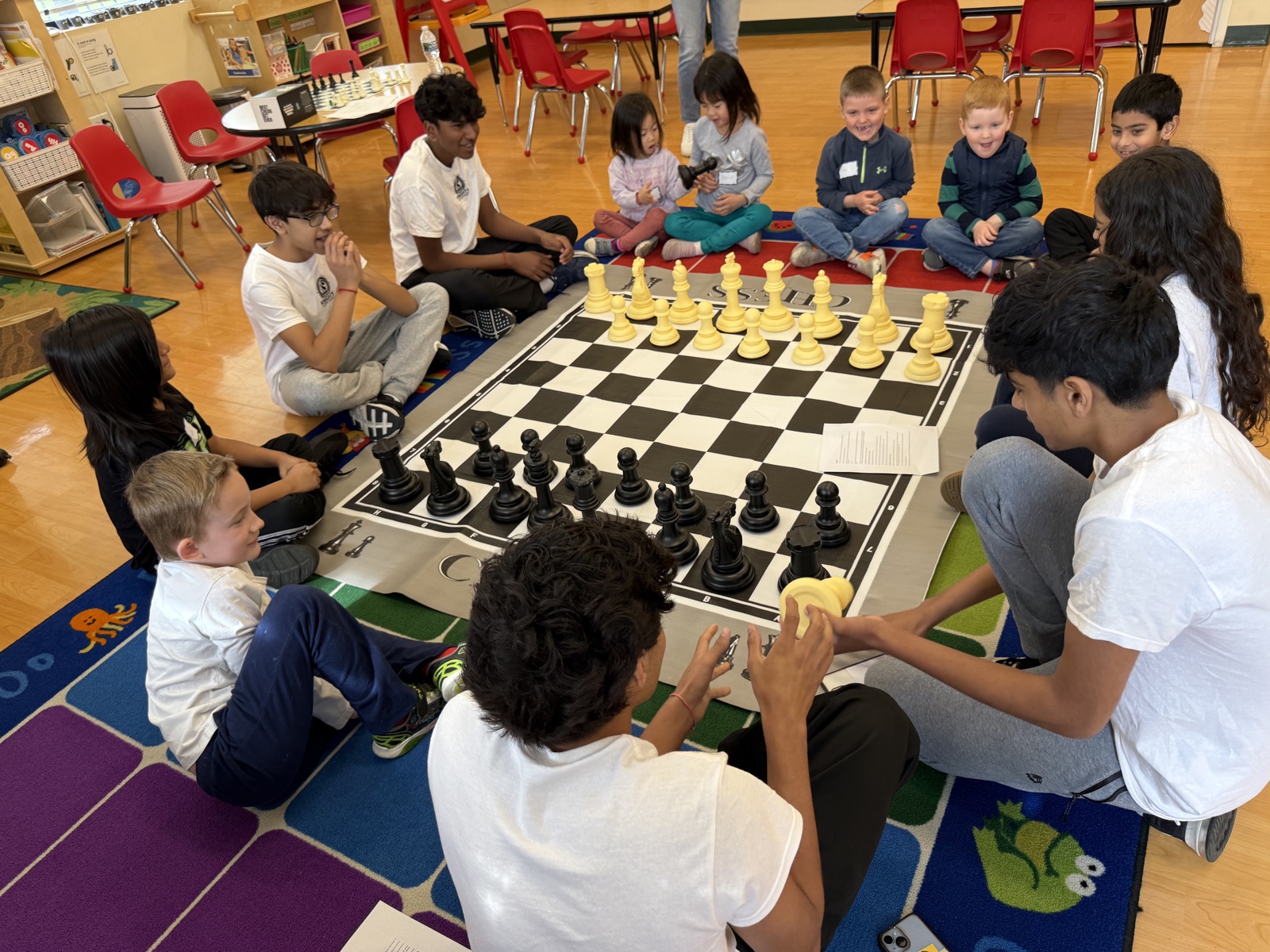 Young children and teenage mentors sitting on a classroom rug around an oversized chessboard, mid-lesson.