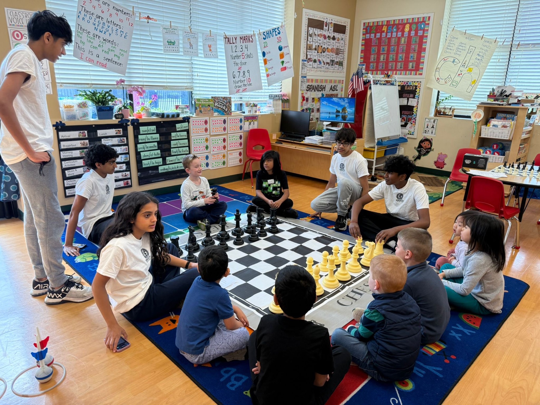 Chess mentors in matching Knights of the Future shirts teaching a group of young children around an oversized chessboard.