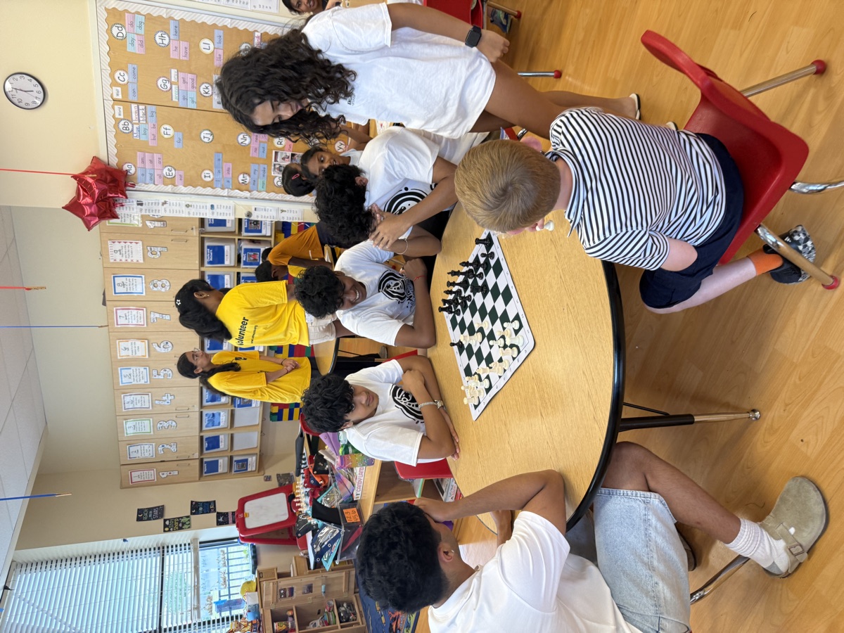 Young children and mentors playing chess around a round table in a classroom.